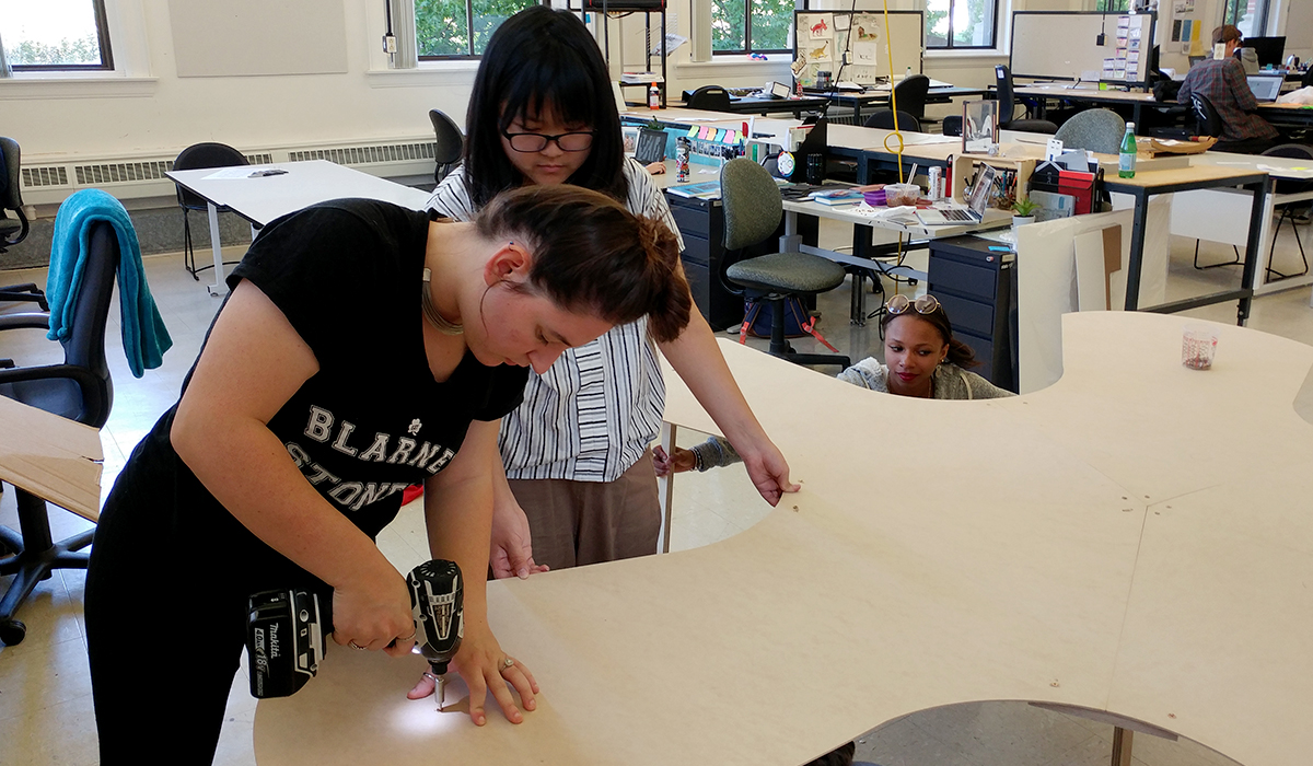 Kha holds odd-shaped board in place while Rosenthal uses electric drill.